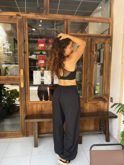 A woman with curly brown hair is standing in front of a wooden storefront in Phu Thuy Ward, Vietnam. She is wearing a black sports bra and black wide-leg pants. Her right arm is raised and her hand is in her hair. The storefront has large glass windows displaying a menu of coffee drinks and prices, including "Espresso 25k", "Americano 25k", and "Latte 40k". Red bags of coffee are visible on shelves behind the glass. A wooden bench is situated in front of the storefront. The ground is paved with light-colored tiles. A plant is visible to the right of the storefront.