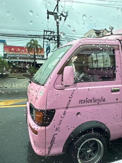 A pink taxi, likely a tuk-tuk or similar open-air vehicle, is parked on a street in Chalong, Thailand. The vehicle is adorned with decorative decals. Water droplets are visible on the windshield, indicating that it is raining or has recently rained. Through the windshield, a busy street scene can be observed, with utility poles and wires overhead, as well as buildings and signage, including "California Fresh Mexican Grill" and "Lamina Films." The sky appears overcast. The text "เขตจังหวัดภูเก็ต" which translates to "Phuket Province Area" is visible on the side of the taxi, confirming the location. Another sign on the taxi reads "TAXI." The overall impression is a candid street-level view of transportation in a tropical, rainy environment.