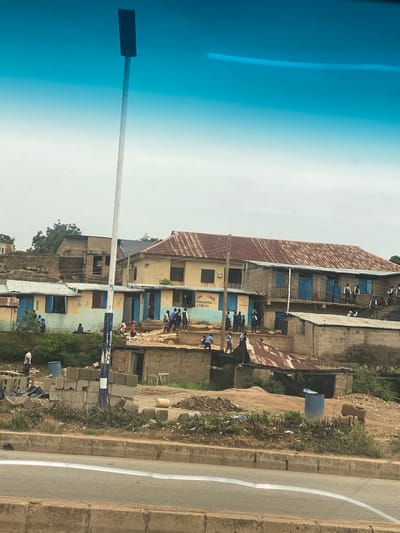 The image depicts a street scene in Barakin Lerawi, Nigeria, under an overcast sky. In the foreground, a concrete road barrier is visible, with a highway running alongside it. Beyond the barrier, a steep, unpaved incline rises, covered in debris, construction materials such as concrete blocks and barrels, and sparse vegetation. Several people, some dressed in light blue shirts and dark pants, and others in casual attire, are scattered throughout the scene.
Dominating the midground and background are several buildings of varying construction and condition. A prominent two-story building with a rusted corrugated metal roof and blue-shuttered windows stands centrally. Another yellow building with a similar roof is to its left, featuring a sign that reads "DAV CHRIS FOUNDATION COLLEGE". To the far left, a partially constructed brick building with an open roof structure is visible. The overall impression is one of a developing or transitional urban environment with active construction and a mix of older and newer structures.