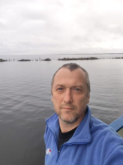 A middle-aged man is captured in a selfie, looking directly at the camera with a neutral and calm expression. He has blue eyes, a short beard, and greyish hair. He is wearing a blue fleece jacket with a stylized "NR" or similar logo in white and red on the left chest, over a black t-shirt. A blue railing or structure is partially visible on the right side of the frame, suggesting he is on a boat, pier, or bridge.

The setting is outdoors, at what appears to be a large body of water, possibly a lake or bay, under a heavily overcast, light grey sky. The water is calm and reflects the muted tones of the sky. Stretching across the water in the mid-ground are several lines of distinct structures. These appear to be remnants of man-made constructions, possibly an old breakwater or defensive line, composed of irregularly shaped rocks and weathered wooden posts or piles that are partially submerged. In the far distance, a faint, dark line indicates a distant shoreline or landmass. The overall weather is gloomy due to the thick cloud cover, creating a serene yet somewhat stark atmosphere with a muted color palette dominated by greys and blues. The location is Medvezhyegorsk, Russia.