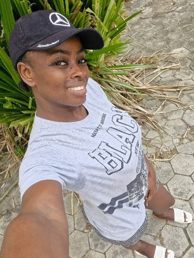 A woman poses for a selfie, wearing a gray t-shirt with "BLACK" printed on it, a black baseball cap adorned with a white Mercedes-Benz logo, and white sandals. She stands on a stone walkway, with green plants and dry grass surrounding her. She smiles slightly, looking directly at the camera. The image is taken from a high angle.