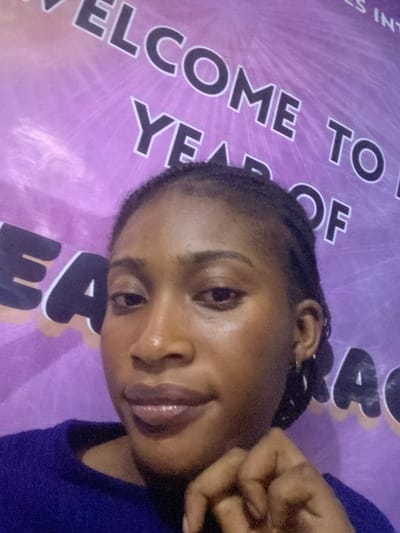 A young adult female, identified as being in Kubwa, Nigeria, is depicted in a close-up, chest-up shot against a purple banner backdrop. She has a dark complexion, dark eyes, and dark, possibly braided hair pulled back from her face. Her lips appear full with a glossy finish. She is wearing a dark blue long-sleeved top and small hoop earrings. Her left hand is positioned near her chin, with fingers slightly curled. The background banner is purple with radiating lines and features large, dark letters outlined in white. Visible text fragments include "WELCOME TO," "YEAR OF," and "GRACE," with additional partially visible letters "ES INT" in the upper right.