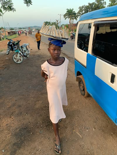 In Gbeleko, Nigeria, a person with a blue headscarf is standing on a dirt road, carrying a load of goods on their head. The person is wearing a light pink dress with a black trim. A blue and white van is partially visible in the upper right of the frame. Two motorcycles are parked on the road. In the background, other people are present, along with buildings and trees under a hazy sky.