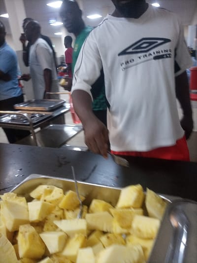 A crowded canteen setting with several individuals lining up. A man in a white and black Umbro Pro Training shirt is using a fork to serve himself pineapple chunks from a large metal tray. The focus is on the tray filled with the yellow fruit. Other people are visible in the background, some holding plates and engaged in conversation. Metal serving trays and carts are also present.