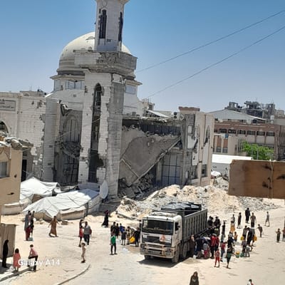 The image depicts a street scene in Khan Yunis, Palestinian Territories, characterized by destruction and humanitarian aid efforts. A large, heavily damaged building, possibly a mosque with a dome and minaret, stands in the background. Its facade is fractured, with debris scattered around its base.
In the foreground, a white Volvo truck with a logo that appears to be for Doctors Without Borders (MSF) is parked on a dusty, sandy street. A crowd of people, including men, women, and children, are gathered around the truck and in the surrounding area. Some individuals are holding yellow jerrycans, suggesting they are collecting or distributing water or other supplies. Several white tents are set up in the middle ground, indicating a temporary settlement or aid distribution point.
The environment appears arid, with a clear blue sky overhead. The presence of damaged buildings, tents, and a gathering of people with water containers points to a situation of crisis and the provision of essential aid in a war-affected area. The image captures a moment of human activity amidst widespread destruction.