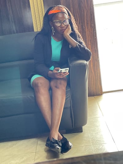 A full shot shows a Black woman sitting on a dark gray couch in what appears to be a waiting area. She is wearing a turquoise dress, a black blazer, a headband with an orange and brown patterned design, and large square-framed glasses. Her right arm is resting on the back of the couch, with her chin propped on her hand. She is holding a white smartphone in her left hand. Her legs are crossed at the knees, and she is wearing black ballet flats with bows. The background consists of a wood-paneled wall to her right and a large window with bright light streaming through to her right. The floor is tiled in a light beige color. The woman's hair is long and black, with some strands falling over her shoulders. The overall impression is one of a professional or business setting. The location is the city of Kwamba, Nigeria.