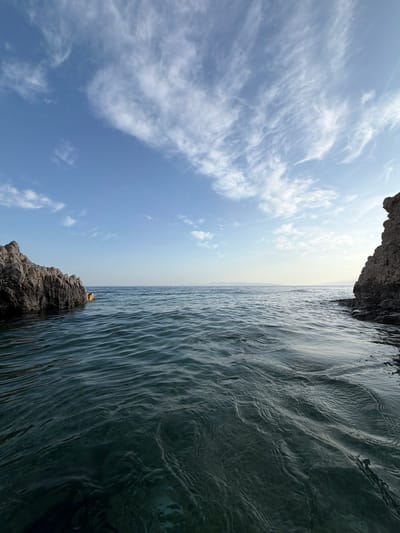 The image displays a serene coastal landscape under a bright daytime sky. The scene is dominated by a clear blue sky and a calm body of water, likely the sea, separated by a distinct horizon line.
The upper portion of the image features an expanse of blue sky, transitioning from a deeper hue at the top to a lighter, almost hazy white-blue closer to the horizon. Scattered across the sky are thin, wispy white clouds, appearing as streaks and patches, indicating fair weather.
The lower half of the image is filled with dark, deep blue-green water, which appears calm with subtle ripples and textured waves reflecting the light. The water extends towards the distant horizon.
Framing the bottom right of the image is a dark, rugged rock formation or cliff edge, showcasing a rough texture. Closer to the center, a smaller, weathered rock outcrop emerges from the water near the horizon. Perched on this smaller rock is a small, indistinct golden or yellowish object, which appears reflective against the darker surroundings.
No human activity or specific event is taking place, presenting a natural and peaceful scene. The time of day appears to be sunny daytime with clear weather. The location is a coastal area, consistent with Grad Rijeka, Croatia. There is no visible text in the image.
