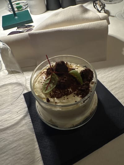 The image displays a dining table setting within a restaurant located in Bolzano, Italy. A clear glass container holds a dessert, consisting of a light-colored creamy base topped with dark chocolate crumbles, three spherical dark chocolate pieces, and two small green leaves with reddish veins, presented on a dark blue paper napkin. To the left, an inverted empty clear glass rests on the white, textured tablecloth. In the background, from left to right, a green stand-like object is partially visible, alongside a clear drinking glass containing liquid and a green item, followed by a white cylindrical container, a folded off-white cloth napkin, two dark cylindrical shakers, and a pair of eyeglasses with dark frames. The scene is static, devoid of people or direct action.