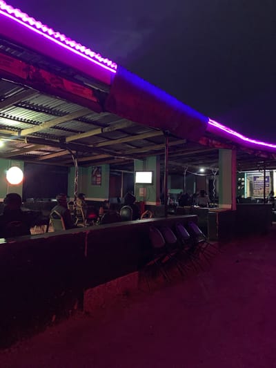 The image depicts an outdoor seating area of a establishment, likely a bar or restaurant, at night. Purple LED lights illuminate the corrugated metal roof structure. Several people are seated at tables and on chairs, some appearing to be engaged in conversation or watching a screen. The environment appears somewhat rustic, with visible wooden beams supporting the roof and a rough, dirt ground surface. There are signs visible inside the establishment, including one that reads "Kitchen". The scene is situated in New Stadium, Nigeria.