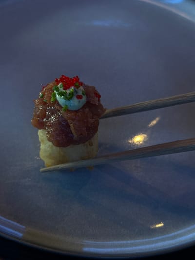In Podgorica, Montenegro, a single piece of sushi, topped with minced tuna, a dollop of cream, red roe, and chives, is being held by two chopsticks over a blue plate. The lighting is dim, with reflections of a bright light source visible on the plate. The sushi appears to be on a light-colored base, possibly rice or tempura.