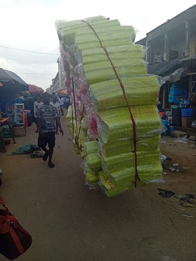 A street-level photograph captures a busy market scene in Ura Nta Umuarandu, Nigeria, under an overcast sky. The prominent subject in the mid-ground is a large, vertically stacked bundle of light-green plastic or paper bowls/containers, each individually wrapped in clear plastic and secured with red ropes. This bundle appears to be in transit, possibly carried by an individual positioned behind it, out of frame. To the left of this bundle, a male pedestrian in a patterned short-sleeved shirt, dark shorts, and sandals walks leftward. Several other indistinct figures are visible further down the street. The ground consists of an unpaved dirt surface with scattered debris. On the left, market stalls with dark canopies display goods including plastic containers and a cooler. On the right, older multi-story buildings with balconies and corrugated metal elements line the street, with plastic buckets and basins displayed outside. A reddish-brown and black bag is partially visible in the lower left foreground.