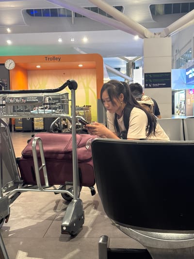 The image displays an indoor scene within an airport terminal, identified as being in Sepang, Malaysia, likely Kuala Lumpur International Airport (KLIA) or KLIA2, based on visible signage in Malay for "Pelepasan Dalam Negeri" (Domestic Departure), "Pelepasan Antarabangsa" (International Departure), and "Ruang Menunggu" (Waiting Area).

In the mid-ground, a young East Asian female, appearing to be in her late teens or early twenties, is seated on a grey upholstered airport bench. She possesses long, dark hair tied into a ponytail with bangs framing her face, and wears a light-colored short-sleeved t-shirt and a dark backpack over her right shoulder. She is observed actively using a mobile phone held in both hands, her gaze directed downwards at the device. A small earring is visible in her left ear. To her right and partially obscured, the back of a male individual with a beige backpack is visible, also seated.

The left foreground is dominated by a silver-colored metal airport luggage trolley with black handles. A maroon soft-sided suitcase rests on the trolley's lower platform. A sign attached to the trolley's upper basket provides information including a "Maximum Load Weight 80 kg" in both Malay and English. A second trolley is partially visible further in the background on the left.

An orange cuboidal structure in the background features the word "Trolley" in white capital letters and a round analog clock displaying approximately 10:10. The background of this structure is decorated with a stylized light green and orange graphic resembling trees. The environment is brightly lit by various artificial overhead fixtures, and the floor is composed of light-colored tiles. White structural pillars and beams are evident, and blurred outlines of retail establishments are visible in the far background.