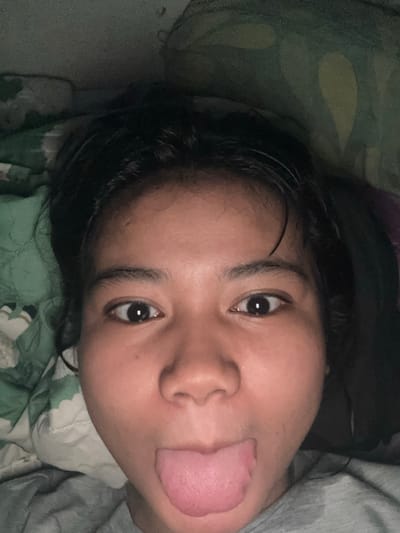 The image shows a close-up, selfie-style shot of a young person's face, with their tongue sticking out. The person has dark hair and is wearing a grey t-shirt. They are lying down, as indicated by the pillows and bedding visible in the background, which appear to be green and white patterned. The lighting is dim, suggesting the photo was taken indoors, possibly at night or in a low-light environment. The context of North Jakarta, Indonesia, does not provide specific details about the scene itself, as it is a general geographical location and does not offer visual cues within the image.