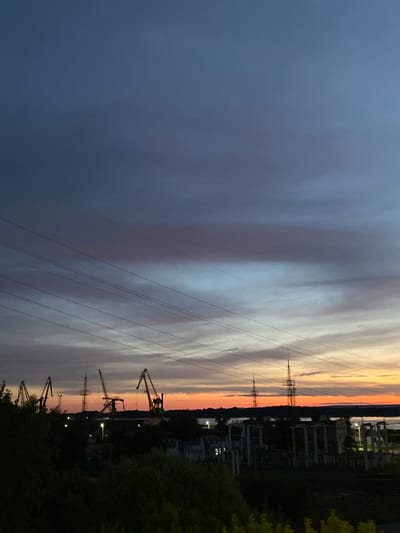 This image depicts a dusky sky over an industrial setting in Novokuybyshevsk, Russia. The sky exhibits a gradient of colors, transitioning from a deep blue on the left to soft oranges and yellows on the right, indicative of either sunrise or sunset. Wispy clouds stretch across the horizon, contributing to the atmospheric effect.
Dominating the foreground are several power lines, stretching diagonally across the frame. In the background, an industrial landscape is visible. This includes multiple large cranes, silhouetted against the colorful sky, suggesting port or construction activity. Scattered lights can be seen within the industrial complex, implying it is either operational or has some lighting during the twilight hours. There is also a transmission tower visible in the distance. Dense foliage, likely trees, frames the right side of the image, adding a natural element to the otherwise industrial scene.
The overall mood is calm and somewhat melancholic, amplified by the dim lighting of dusk. The scene is devoid of people. No visible text is present in the image.