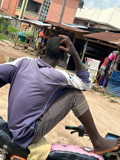 A man is sitting on a motorcycle, facing away from the camera, with his right hand on his head. He is wearing a purple and white long-sleeved shirt and grey pants. The motorcycle has a black seat and a pink gas tank. In the background, there are several market stalls with corrugated metal roofs. One stall displays colorful items hanging from the roof. Another stall has clothing racks with various garments. The ground is sandy and appears wet in some areas, suggesting recent rain. Buildings with orange and beige facades are visible further in the background. The overall scene depicts a street market environment in Ihiagwa, Nigeria.