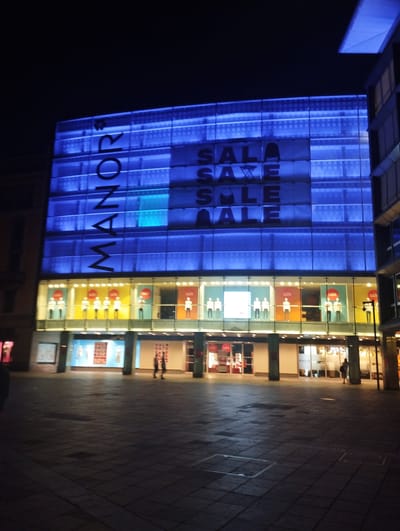 The image shows a department store at night, illuminated by bright blue lights on its upper facade. Large text, also in blue, spells out "MANOR SALE SALE SALE" across the building's front. The lower level of the store features shop windows displaying mannequins and promotional material with the word "Sale" visible. Several people are walking on the tiled pavement in front of the store. The scene appears to be a public square in a city, consistent with the location of Manor department store in Lugano, Switzerland.