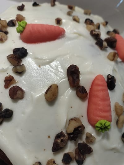 The image shows a close-up view of a carrot cake. The cake is covered in white frosting and decorated with chopped walnuts and two marzipan carrots. The carrots are orange with green frosting tops. The cake appears to be on a white plate or surface. The setting is not specified, and there is no indication of the city of Crauthem, Luxembourg.
