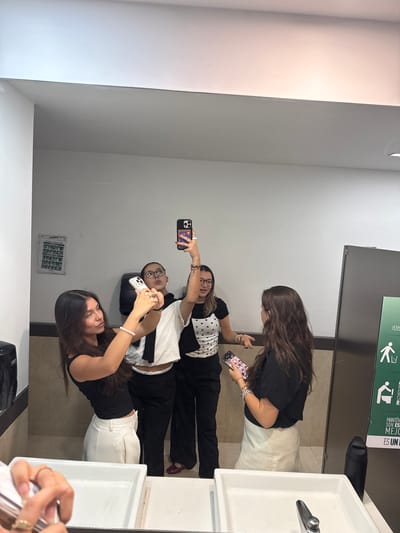 A photo of four women in a bathroom, likely a public restroom, in what can be assumed is San Andrés Cholula, Mexico. The women are taking a selfie in front of a mirror. They are positioned in a line, with the first woman holding up her phone to take the picture. The women are dressed in casual clothing. The restroom has white walls and tile flooring. There are sinks and a mirror visible in the frame. A sign in Spanish is located above the sinks.