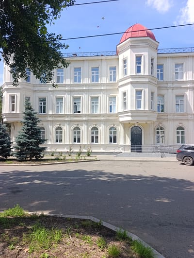 This is a full exterior, eye-level shot of a white, ornate, multi-story building in Orenburg, Russia, on a clear, sunny day. The building has a prominent red domed turret and numerous white-framed windows. In the foreground, to the left of the building, are two tall, blue-green fir trees and to the right is a dark SUV parked on the asphalt road. A tree branch with green leaves hangs from the upper left corner of the frame, obscuring some of the building and the sky. The road in front of the building is grey asphalt, and there is a patch of grass and soil along the curb on the left side. The building appears to be in good condition and is likely a public building or institution.