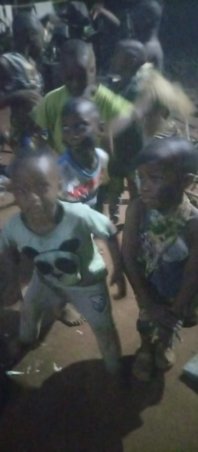 This image captures a group of approximately seven to ten young children gathered in a low-light environment, likely at night, in Amokwue-Affa, Nigeria. The children, appearing to be toddlers to early elementary school age, are seen standing or crouching on an unpaved, dirt-like ground surface.

In the immediate foreground, to the left, a child wears a light-colored t-shirt featuring a prominent panda face graphic and light-colored pants. A faint, illegible logo, possibly including the numbers "198" or "1984" within a shield design, is visible on the right thigh of the pants. This child is barefoot and adopts a kneeling or crouching posture. To their right, another child is visible, also barefoot, wearing a dark, patterned sleeveless top and dark bottoms.

Behind these two children, several others are present. One child wears a light-colored top with a design. Further back, multiple children are dressed in light green or yellowish-green shirts. The faces of many children are obscured by darkness and slight blurring, making individual expressions unclear, but they do not appear to be distressed. One child in the deeper background has a notable light-colored element on their head, possibly light hair or a head covering.

The setting is characterized by very dim illumination, strongly suggesting nighttime outdoors or an indoor space with very poor lighting. The background is indistinct and dark, with vague shapes that could be foliage or structures. No discernible objects other than the children's clothing are visible. The overall scene depicts a casual gathering or interaction among children in a rural or community setting.