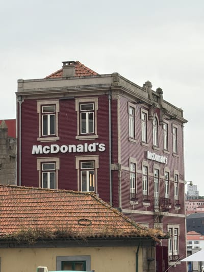 This is a vertical, eye-level medium shot of a historic building with a McDonald's sign prominently displayed. The building features a dark red, corrugated facade with numerous windows. White, block-lettered "McDonald's" signage is affixed vertically to the building's face, with the "M" near the top and the "s" near the bottom. The architecture suggests an older European style, with decorative stone accents around the windows and along the roofline. The roof itself is covered in red tiles. To the right of the main building, a portion of an adjacent building with a yellow facade and a green-framed window is visible. The sky is overcast, suggesting a cloudy or possibly rainy day. The overall impression is that of a McDonald's restaurant integrated into the architecture of a historical European city, likely Porto, Portugal, given the context. There is no visible activity or people in the frame. The lighting is diffused due to the cloudy weather.
