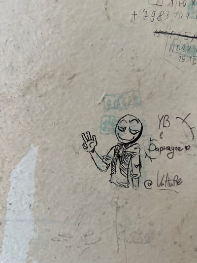 The image displays a section of a textured, light-colored wall, possibly concrete or plaster, marked with various graffiti. In the center-left, a cartoonish drawing of a figure with a wide smile and closed eyes is depicted. The figure wears a hooded jacket and holds its right hand up in a peace sign. To the right of the drawing, graffiti in Russian script is visible, including "УВ в Барнауле" which translates to "UV in Barnaul," and "@ Vatture." Above these writings, other inscriptions are present, some of which are numbers like "+7983104" and "19LE." The overall environment appears to be an outdoor urban setting, likely a public wall in the city of Barnaul, Russia, covered in various forms of street art and inscriptions.