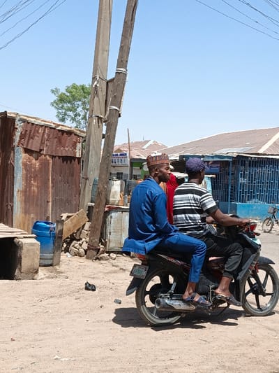 An outdoor scene in Gwagwarwa, Nigeria, depicts two males seated on a black underbone motorcycle on an unpaved, dusty reddish-brown dirt road under a clear blue sky. The motorcycle is stationary. The driver, wearing a blue baseball cap, a black and white striped short-sleeve shirt, and dark pants, occupies the front seat, his feet in dark sandals on the footpegs. A passenger sits behind him, dressed in a blue long-sleeve tunic, matching blue pants, and a multi-colored patterned cap, his feet in blue sandals also on the footpegs. A third individual, partially obscured between the driver and passenger, wears red clothing. To the left, a structure composed of rusted corrugated metal sheets stands next to a blue plastic barrel. Two utility poles, one concrete and one wooden, are strapped together, rising from the left mid-ground, with overhead wires visible. In the background, various buildings with corrugated metal roofs are visible, including one with a blue metal grate facade. Solar panels are mounted on some roofs. A green tree is visible in the distant background to the left. A bicycle is parked further down the road to the right.