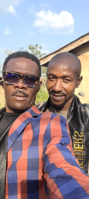 Two adult males are depicted in a selfie taken outdoors in Alayi, Nigeria, under a partly cloudy sky. The individual positioned on the left, partially obscured by the foreground, wears dark rectangular sunglasses with a mottled frame, a mustache, and a black shirt visible beneath a red and blue plaid long-sleeved shirt. The individual on the right, smiling and displaying teeth, has a shaved head, a short beard, and wears a light brown collared shirt. A black sash or stole with yellow lettering, partially visible as "EDEK?", is draped over his left shoulder. Both individuals are looking directly at the camera. The background on the left features green foliage, while the right displays a building with a light brown wall, a dark roofline, and a small, reflective object attached to the wall.