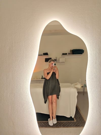 A woman is taking a selfie in a spa located in Vinnytsia, Ukraine. She is standing in front of a mirror with integrated lighting. She is wearing a dark grey towel wrapped around her body and a clear plastic shower cap on her head. She is holding a smartphone in her right hand, capturing the reflection. In the background, a massage table covered with white linens is visible, along with shelves displaying towels and bottles. The room appears to be a treatment room within a spa.