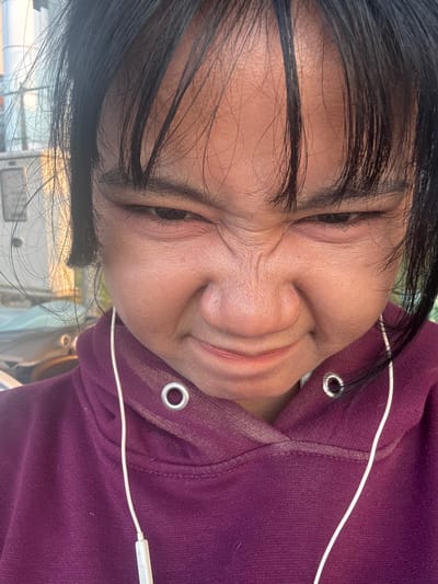 A close-up, low-angle, head-and-shoulders shot captures a young person with dark hair and a maroon hoodie, looking directly at the camera with a scrunched-up, annoyed expression. Their brow is furrowed, eyes are narrowed, and the corners of their mouth are turned down. White earbuds are visible, with one wire trailing down the front of the hoodie. The background is out of focus, showing a blurry outdoor scene with some vertical structures that appear to be in a city environment, possibly in North Jakarta, Indonesia. The lighting suggests it might be daytime, with sunlight casting shadows and highlights on the person's face.