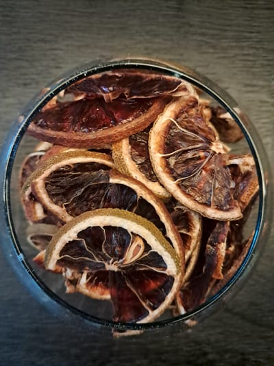 A top-down, close-up shot shows a clear glass bowl filled with dried citrus slices. The slices, likely oranges or grapefruits, are arranged haphazardly, filling the bowl. The fruit's flesh appears dark brown and shriveled, while the rinds are a lighter tan with some green. The bowl is sitting on a dark gray surface. The exact location within Vicente López, Argentina, cannot be determined from the image.