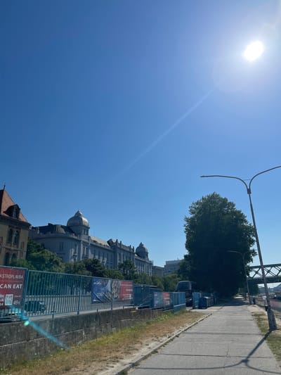 This is a daytime, eye-level shot of a city street in Bratislava, Slovakia, looking towards the Danube River. The sun is bright and high in the sky, casting lens flare and glares. The street is paved with concrete and lined with a metal fence. Behind the fence, there are several buildings, including a light-colored building with domes and an older building with a red tile roof. There are also trees, a lamppost, and a bridge in the distance. Several advertisements are visible on the fence. A few vehicles, including a bus, are parked on the street. The general impression is one of a modern urban environment.