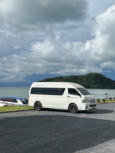 A white van is parked on a gravel lot next to a body of water. The van has tinted windows and text written on its side in Thai. Several boats are docked in the water behind the van. In the distance, a lush, green island rises from the water. The sky is filled with dramatic, gray clouds. The scene appears to be in Ratsada, Thailand, a coastal area known for its beautiful scenery and boat tours.