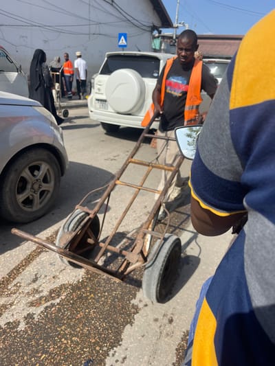 An urban street scene in Zanzibar, Tanzania, is depicted under direct sunlight. In the midground, a Black male wearing a black t-shirt, an orange safety vest, and light-colored pants is pulling a rusty, four-wheeled hand cart forward. The cart is empty. To the immediate left of the cart is the front wheel and lower portion of a silver or light-colored vehicle. Behind the man with the cart, a white Toyota Rav4 SUV is parked, identifiable by its rear-mounted spare tire and a partially visible Tanzanian license plate "T 933 LFG". In the background, on the left side of the street, several individuals are visible: one person in a black abaya, another in an orange vest, and a third in a white shirt and head covering, among others. Buildings line the street, and overhead power or communication cables are present. A blue road sign with a white pedestrian icon is mounted on a pole above the white SUV. The foreground on the right shows the shoulder and arm of an individual wearing a blue and yellow striped shirt, with a motorcycle mirror reflecting the sky and trees. The asphalt road surface exhibits dark, wet-looking patches.