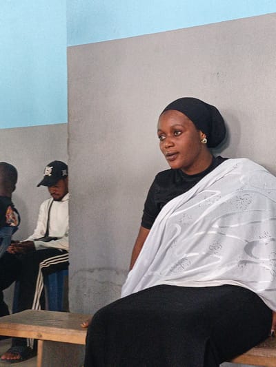 A medium shot shows a woman seated on a wooden bench in what appears to be an interior room. The woman has dark skin and is wearing a black head covering, a black top, and a long black skirt or dress. A white shawl or garment is draped over her shoulders. She is looking to her left, away from the camera.
In the background, partially obscured, are two other individuals. One is wearing a black baseball cap and a white jacket, and the other is wearing a dark garment. They are also seated. The room has walls painted in a light blue and gray color scheme. The location is in the city of Fagge C, Nigeria.