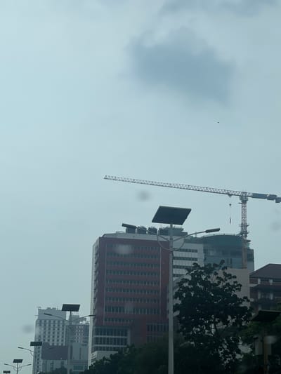 The image depicts a view of a city skyline in West Jakarta, Indonesia, characterized by overcast skies and the presence of construction. In the foreground, a light pole with solar panels is visible, along with some trees partially obscuring the view of buildings behind it. The most prominent structures are several multi-story buildings, including one with a red facade and rows of windows, and another under construction featuring a large crane extending across the upper portion of the frame. The sky is a pale gray, with a large, diffused cloud formation and a small, dark object, possibly a bird or a drone, in the distance. The overall scene suggests an urban environment undergoing development under cloudy weather conditions.
