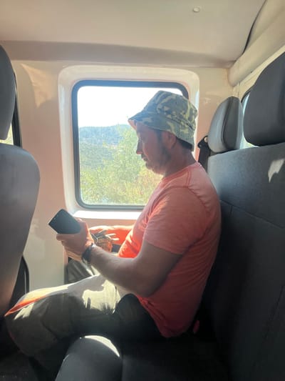 A male individual is observed seated within a vehicle, located in or near Carrazeda de Ansiães, Portugal. The subject, viewed in profile facing left, wears a camouflage bucket hat, an orange short-sleeved t-shirt, and dark-colored trousers. He holds a black smartphone in his left hand, while his right hand manipulates a smaller, dark object. A dark-colored smartwatch is present on his left wrist. The vehicle's interior features dark gray padded seats with headrests and a light-colored upper interior. Through a rectangular window to the left of the subject, a brightly lit outdoor scene is visible, comprising green foliage, distant hills, and a clear sky. Sunlight streams through the window, creating distinct shadow patterns on the subject's lower body and the adjacent seat.