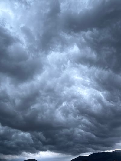The image displays a dramatic, heavily clouded sky extending across most of the frame, with a dark mountain silhouette visible along the right edge. The sky is dominated by dense, turbulent masses of dark grey and bluish-grey clouds. Patches of brighter, diffused light are visible where the cloud cover is less thick or where light is breaking through, creating a striking contrast and highlighting the voluminous, undulating forms of the clouds.
Towards the right side of the vertically-oriented image, a stark, dark silhouette of a rugged mountain or cliff face is clearly visible, its jagged profile outlined against the brighter, hazy areas of the sky. Some sections of the cloudscape, particularly near the mountainous edge, exhibit distinct wavy or layered patterns, indicating dynamic atmospheric movement.
No people, man-made objects, or visible text are present. The scene evokes a sense of awe and powerful natural forces, suggesting heavy overcast weather, possibly an impending or ongoing storm. The general lighting suggests it could be an overcast day, late afternoon, or early evening. The mountainous terrain is consistent with the provided location context of Podgorica, Montenegro.
