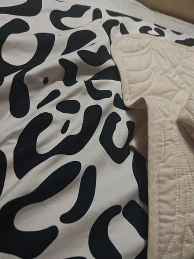 The image shows a close-up view of a beige quilted blanket folded over a black and white patterned fabric. The pattern on the fabric appears to be abstract or organic shapes. The scene is indoors, likely in a bedroom, as suggested by the presence of bedding. There are no people visible in the image. The lighting is soft and diffused. The location in Kokshetau, Kazakhstan is not discernible from the image.
