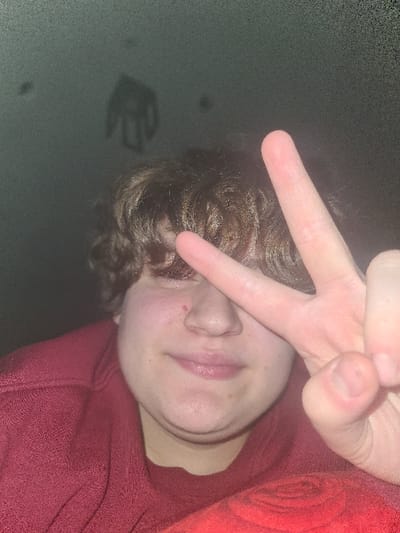 A close-up depicts a person partially obscured by a hand making a "peace" sign across their face. The individual has fair skin, brown curly hair, and is wearing a red garment. The hand appears pale, and the focus is primarily on the face and hand. The background is a dark, blurry wall with a shadow of a hand cast on it.