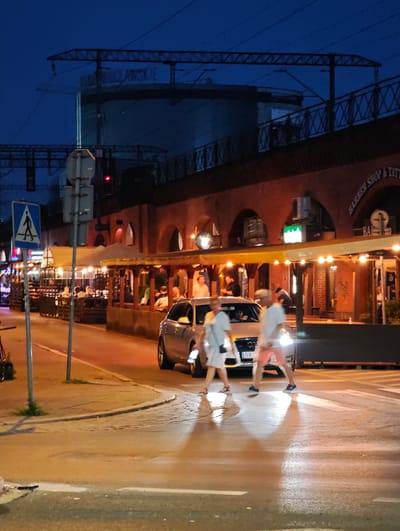 The image shows a street scene at dusk in Wrocław, Poland. A modern silver Audi A4 is stopped at a pedestrian crossing, with its headlights illuminated. Two individuals are walking across the street, one in a white dress and the other in shorts and a t-shirt. Behind the pedestrian crossing, an outdoor dining area is visible, with people seated at tables under awnings lit by string lights. The buildings in the background appear to be old brick structures with arched entrances. Above the buildings, a bridge or overpass with train tracks is visible, and further in the distance, a large cylindrical structure with text that reads "WROCŁAWSKIE" can be seen. The street is wet, reflecting the lights.