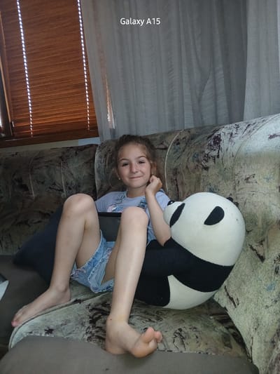 The image captures a young girl lying sideways on a patterned couch, smiling at the camera. Her head is positioned towards the left of the frame, and her legs extend to the right. She has light brown hair and fair skin, and is wearing a light-colored t-shirt and light blue denim shorts. Alongside her, a large, black and white panda plush toy rests on the couch, its head near her right arm. The couch features an abstract or faded floral pattern in shades of green, brown, and grey.
In the background, to the left of the girl's head, a window is partially covered by sheer white curtains and horizontal wooden blinds, allowing bright streaks of daylight to enter the room. The setting appears to be indoors, likely a living room or bedroom in a domestic environment. The girl's cheerful smile conveys a sense of happiness and relaxation. Along the far-left edge of the image, the text "Galaxy A15" is visible vertically. The scene takes place in Obrochishte, Bulgaria, during the daytime.