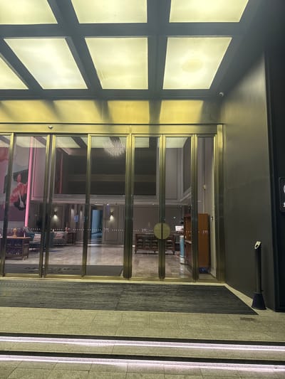 The image shows the entrance to the Mercure Hotel in Huangpu District, China, at night. The entrance features large glass sliding doors with gold-colored frames. Above the entrance, a ceiling with translucent panels is illuminated from within. A security camera is visible on the ceiling near the right side of the entrance.
Inside the hotel, visible through the glass doors, is a well-lit lobby with a marble floor. There are seating areas with sofas and tables. A reception desk or counter is also partially visible. In front of the glass doors, there is a black ribbed doormat. The steps leading up to the entrance are made of stone and are illuminated with white lights.
To the right of the entrance, a dark gray wall is visible, with a tall, slender black object that appears to be an outdoor ashtray or waste receptacle. A small white security camera is mounted on the wall above the entrance. Another sign with a logo and text is partially visible on the far right edge of the image. The overall scene suggests a modern, upscale hotel entrance.