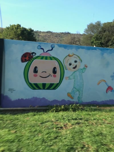The image displays a brightly colored outdoor mural featuring characters from the children's show CoComelon, set against a backdrop of natural scenery. The photograph itself is rotated 90 degrees clockwise, showing the sky and trees on the left and grass on the right.
The central part of the mural features a large, smiling character head with a distinctive green and black watermelon-patterned hat, light pink skin, and wide black eyes. To the left of this character is a red ladybug with black spots. Above and to the right, another depiction of the character, a baby in a light blue, star-patterned onesie, is shown lying on its back with a happy expression. A partially visible toy or rattle with red and yellow parts hangs in the upper right corner of the mural. The mural's background is a light blue with white cloud-like shapes, and a purple stripe runs along its bottom edge. Small, illegible text, likely a signature, is visible near the bottom right of the watermelon character.
Beyond the mural, a clear blue sky is visible, indicating a sunny daytime. A hillside covered in trees and green foliage stretches across the horizon. A thin, dark pole or antenna stands against the sky. To the right of the mural, a patch of green grass suggests an outdoor, possibly roadside, location. The overall scene is cheerful and bright, capturing a whimsical mural in an open-air environment in Germiston, South Africa.