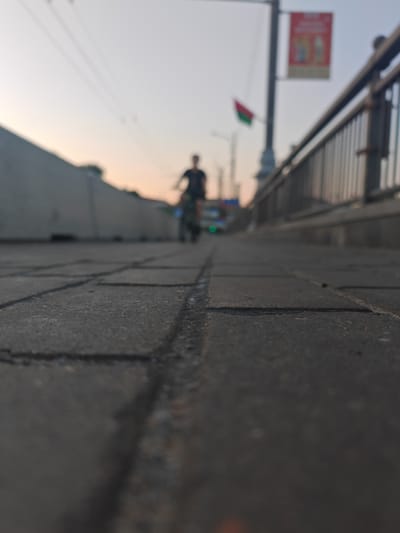 The image depicts a low-angle shot of a paved walkway, likely a sidewalk or pedestrian path. The foreground is dominated by dark gray interlocking pavers, with visible cracks and lines between them. In the background, a person is riding a bicycle. The cyclist is out of focus, appearing as a dark silhouette against the sky. Beyond the cyclist, there's a blurred view of a city street with some vehicles and a traffic light that appears green.
To the right of the frame, a metal railing with vertical balusters runs along an elevated structure, suggesting the walkway might be on a bridge or overpass. Further back, a lamppost is visible with a red banner displaying some text and imagery, although it is too blurred to read. A small flag, tri-colored in red, green, and white, is also attached to the lamppost. The sky is a pale gradient of soft blues and pinks, indicating either sunrise or sunset. The overall lighting suggests it is early morning or late evening. The scene appears to be an urban environment in Mahilyow, Belarus, possibly during fair weather given the outdoor activity. The mood is tranquil, with the focus on the pedestrian environment and a solitary cyclist.