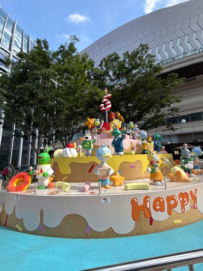 This outdoor shot depicts a multi-layered display of cartoon character figures arranged on a structure resembling a cake, with the word "Happy" prominently displayed. The scene is set in an urban environment, as evidenced by the modern architectural building in the background. Green trees partially obscure the building. A blue sky is visible in the upper left corner. The precise location is Liangjiang, China.