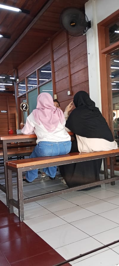 The image shows two women sitting on a bench in a restaurant. The woman on the left is wearing a pink headscarf and blue jeans. The woman on the right is wearing a black headscarf and a beige dress. The bench is made of wood and metal. The floor is tiled with white and red tiles. There is a ceiling fan in the background and a glass window to the right. The restaurant is located in Cangkuang, Indonesia, at the coordinates -7.044 latitude and 107.556 longitude. The women are speaking to each other, and the woman on the left has her hand raised, possibly gesturing.