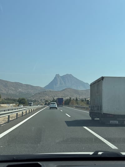 The image shows a view from a vehicle on a highway in Villajoyosa, Spain. The highway is asphalt with clear white lane markings. On the right side of the highway, a white trailer truck is visible, with its rear license plate partially obscured by shadow but showing characters that may be read as "C-694 BKM". On the left side of the highway, there is a metal guardrail. In the middle distance, several vehicles are driving, including a white car and a blue truck. Beyond the highway, the landscape features dry, rolling hills with sparse vegetation and trees. In the background, a large, rugged mountain with a distinctive rocky peak dominates the horizon under a clear blue sky. The overall scene suggests a sunny day on a major road in a mountainous, arid region.