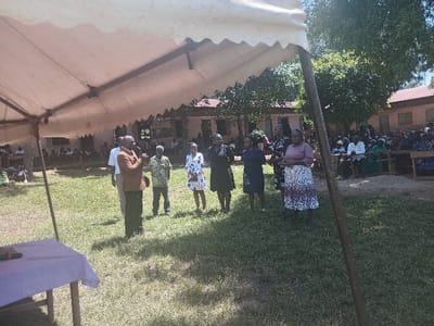 The image depicts an outdoor public gathering in Idakho South ward, Kenya. In the midground, a man wearing a brown jacket over a white shirt is speaking into a dark microphone, standing on a grassy field. He is positioned among approximately eight to ten other adults, including another man in a patterned shirt and several women dressed in various skirts/dresses and blouses. A white canopy tent partially covers the upper left portion of the frame. In the lower left, beneath the tent, a wooden table draped with a light purple cloth holds a black microphone. In the background, a large audience of individuals is seated on benches, facing the group of speakers. Behind the audience, multiple single-story buildings with brown roofs are visible, interspersed with green trees and foliage. The scene is illuminated by natural daylight under a clear sky.