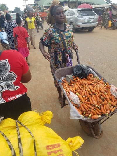 The image depicts a busy street market scene in Nnobi, Nigeria, during daylight hours.
In the midground, a dark-skinned woman with dark hair pulled back, wearing a multi-colored patterned short-sleeved tunic and sandals, is actively pushing a brown wheelbarrow. The wheelbarrow is filled with numerous orange carrots, some loose and some contained in small, clear plastic bags. A black plastic bag is also visible within the wheelbarrow. The woman has a dark bag with a purple and white patterned strap slung across her body.
In the immediate foreground, to the bottom left, several large yellow sacks are visible, partially secured with striped fabric. Red text, including "BEG," is legible on one sack. Behind these sacks, the upper back of a person wearing a red shirt with a white floral pattern is partially visible, facing away from the viewer.
The background reveals a bustling unpaved road. On the left, a light blue scooter is parked. Several pedestrians are walking, including a person in a red t-shirt and pink skirt, another in a black and white patterned dress, and a woman in a yellow t-shirt and light brown pants carrying an object on her head, walking towards the viewer. A man on a motorcycle is further back on the road. Buildings with diverse roofing materials, including corrugated iron, line the street.
On the right side of the background, a silver SUV, appearing to be a Lexus RX model, is either parked or moving slowly. Additional pedestrians are present, one carrying a yellow jerrycan. A red umbrella-like structure is visible above market stalls. The ground throughout the visible street area is dusty, light brown earth. Overhead, power lines are discernible against the sky.